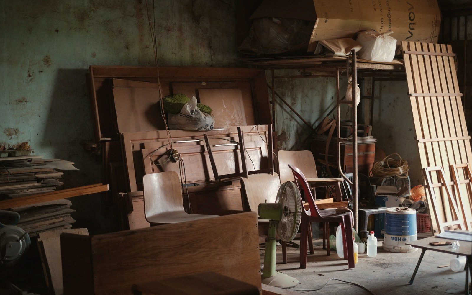 Cluttered storage room with stacked chairs, wood boards, fan, and bottles against peeling walls